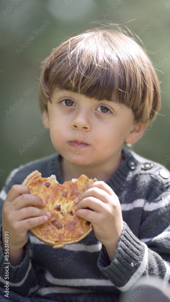 Young boy eating a pizza outdoors, holding the slice with both hands, looking directly at the camera, enjoying his snack during a break in a natural setting with greenery in the background