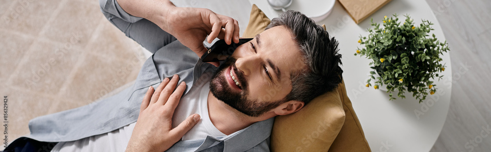 A bearded man in casual attire smiles while working remotely from home, talking on his phone. A bearded man in casual attire smiles while working remotely from home, talking on his phone.