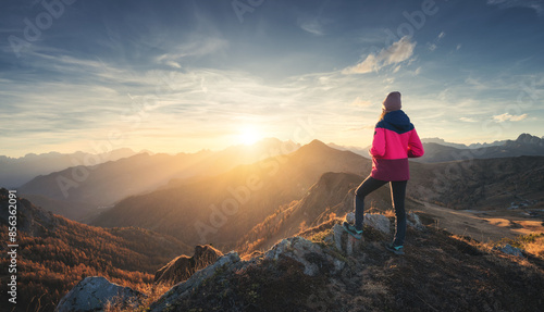 Young woman on mountain peak and beautiful mountain valley in haze at colorful sunset in autumn. Dolomites, Italy. Sporty girl, mountain ridges in fog, orange grass, trees, golden sun in fall. Hiking