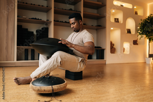 Low-angle view of African young man in trance sitting playing on steel tongue drum by hands on floor at home with cozy interior. Silence calm filling room by sounds of music from musical instrument. photo