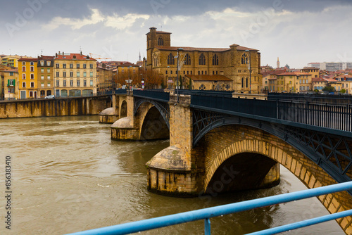 Scenic view across Isere river of Romans-sur-Isere embankment with St. Barnard Roman Catholic Cathedral and ancient stone arched bridge in winter, France