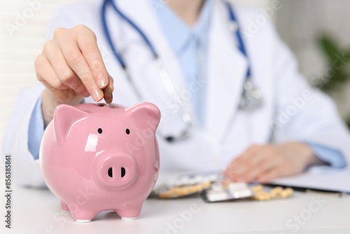 Doctor putting coin into piggy bank at white table indoors, closeup