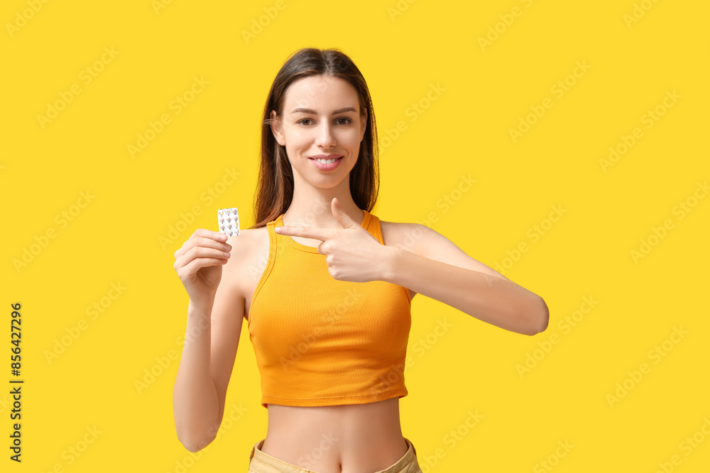 Beautiful young woman pointing at birth control pills on yellow background Beautiful young woman pointing at birth control pills on yellow background