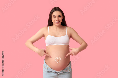 Young pregnant woman with heart made of cream on her belly against pink background