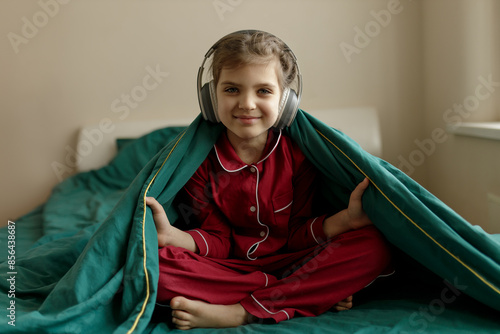 Happy 7 year old girl in red pyjamas and headphones sits in green bed with blanket at home