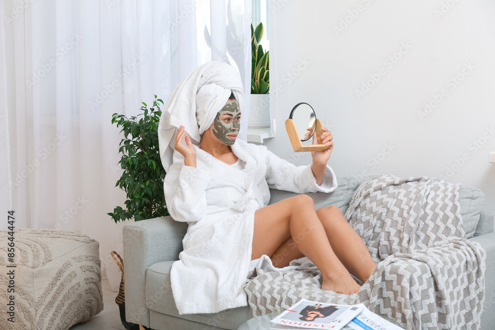 Φωτογραφία Young African-American woman in bathrobe with clay facial mask and mirror at hom