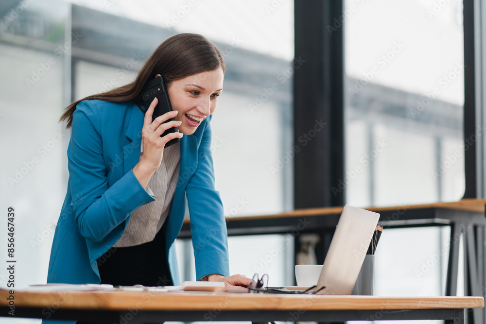 A woman is talking on her cell phone while sitting at a desk. She is wearing a blue jacket and a white shirt A woman is talking on her cell phone while sitting at a desk. She is wearing a blue jacket and a white shirt