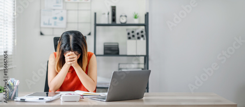 A woman is sitting at a desk with a laptop and a stack of papers. She is looking at the laptop and she is in a state of distress photo