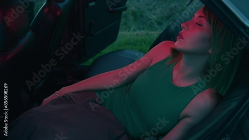 A young stylish woman in a green dress and green wig sits with her eyes closed in a retro car in a red light with flowers all around. photo