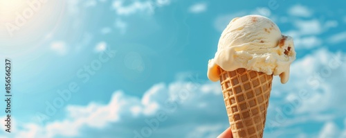 Hand holding a waffle cone ice cream against a blue sky with fluffy white clouds and sunlight. Perfect summer treat for any warm day.