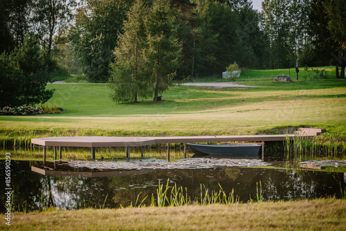 Blome, Latvia - September 11, 2023 - Small dock with a rowboat on a tranquil pond, surrounded by lush grass and trees in a peaceful countryside setting. Copy space. photo