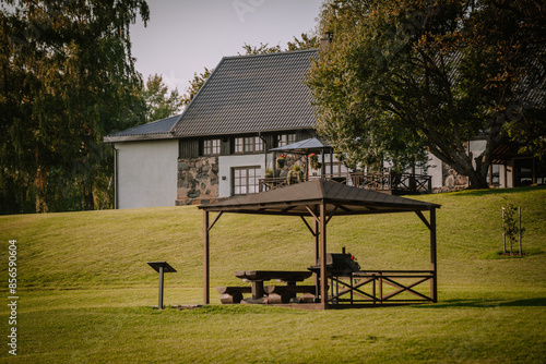 Blome, Latvia - September 11, 2023 - Wooden gazebo with picnic table and grill on a grassy hill, with a rustic house and garden in the background. Copy space.