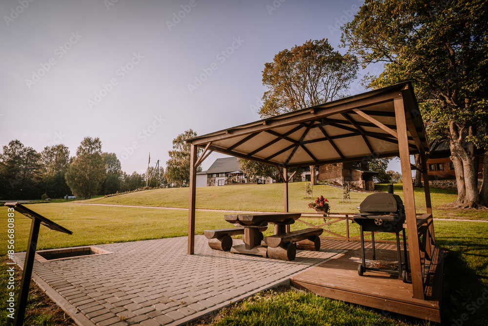 Blome, Latvia - September 11, 2023 - Wooden gazebo with picnic table and grill on a brick patio, surrounded by green grass, trees, and a clear sky in a park setting. Blome, Latvia - September 11, 2023 - Wooden gazebo with picnic table and grill on a brick patio, surrounded by green grass, trees, and a clear sky in a park setting.
