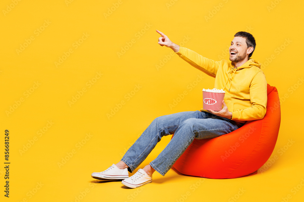 Full body young smiling happy man he wears hoody casual clothes sit in bag chair eat pop corn watch movie film point finger aside isolated on plain yellow orange background studio. Lifestyle concept. Full body young smiling happy man he wears hoody casual clothes sit in bag chair eat pop corn watch movie film point finger aside isolated on plain yellow orange background studio. Lifestyle concept.