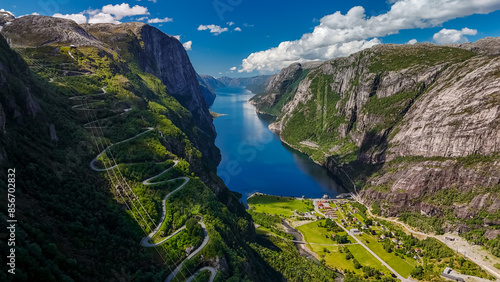 Fotografija An aerial view of a winding road snaking through the lush green mountainsides, overlooking a serene fjord in Norway on a beautiful sunny day