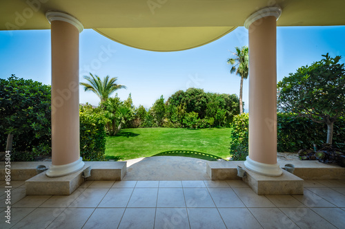 A view from a sunlit terrace framed by elegant columns, overlooking a lush green lawn on a bright summer day. The scene is serene and inviting, with vibrant greenery stretching across the landscape photo