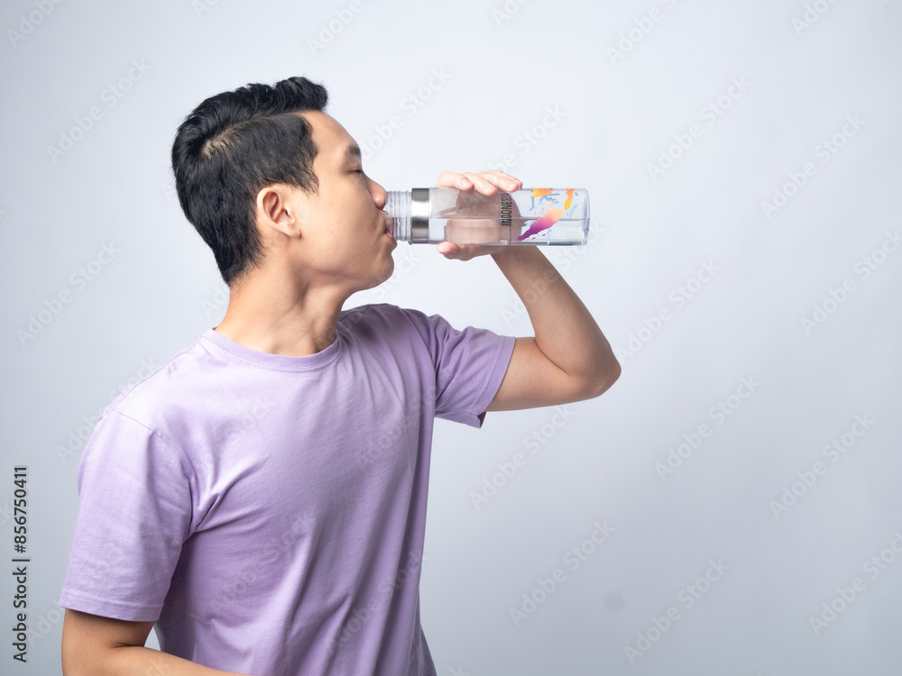 Young Asian man in a light purple shirt, drinking water from a transparent water bottle, standing sideways against a plain light grey background, highlighting hydration and healthy lifestyle. Young Asian man in a light purple shirt, drinking water from a transparent water bottle, standing sideways against a plain light grey background, highlighting hydration and healthy lifestyle.