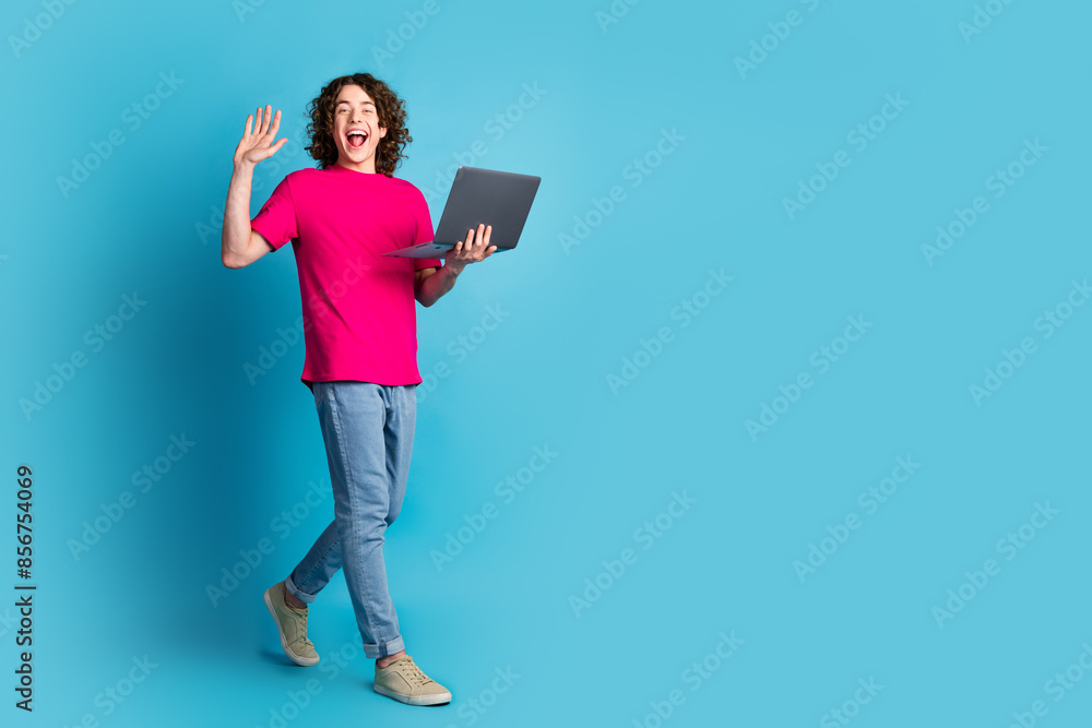 Full size photo of nice young man netbook walk arm wave hi wear t-shirt isolated on blue color background Full size photo of nice young man netbook walk arm wave hi wear t-shirt isolated on blue color background