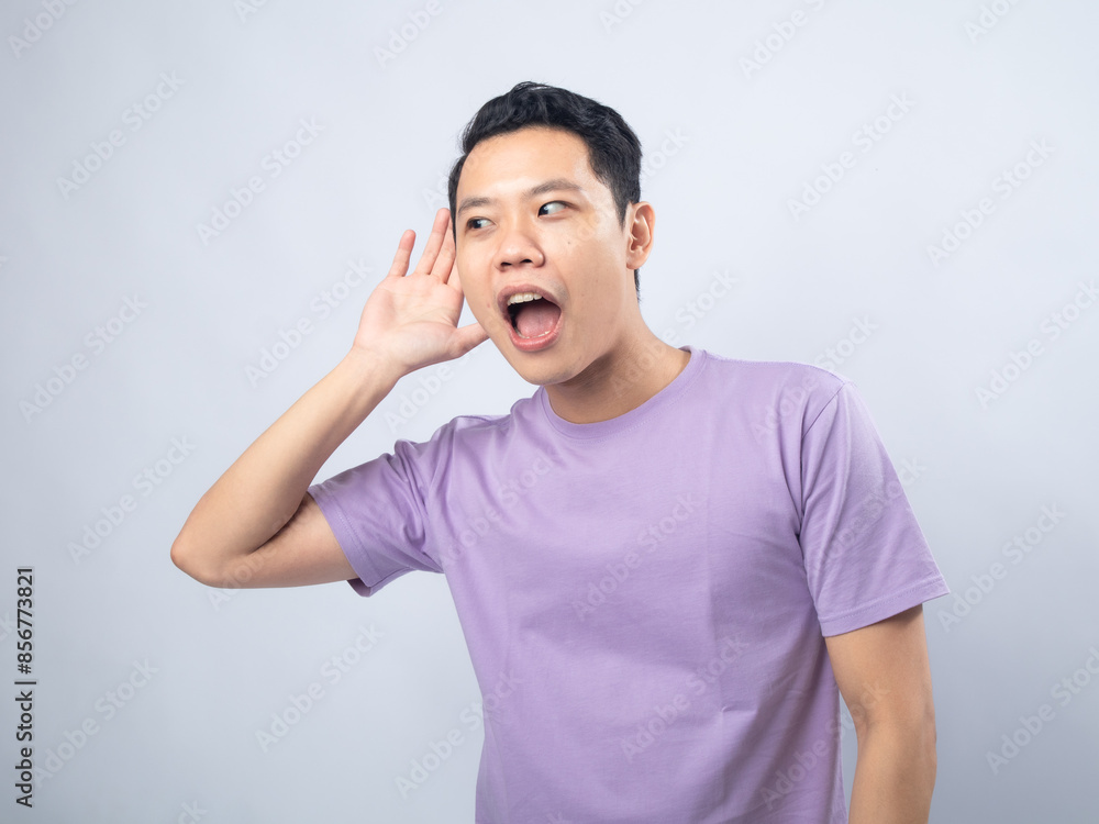 Young Asian man in a lavender t-shirt cupping his ear and looking surprised as if trying to hear something better. Studio shot on a plain background, highlighting his curious expression Young Asian man in a lavender t-shirt cupping his ear and looking surprised as if trying to hear something better. Studio shot on a plain background, highlighting his curious expression