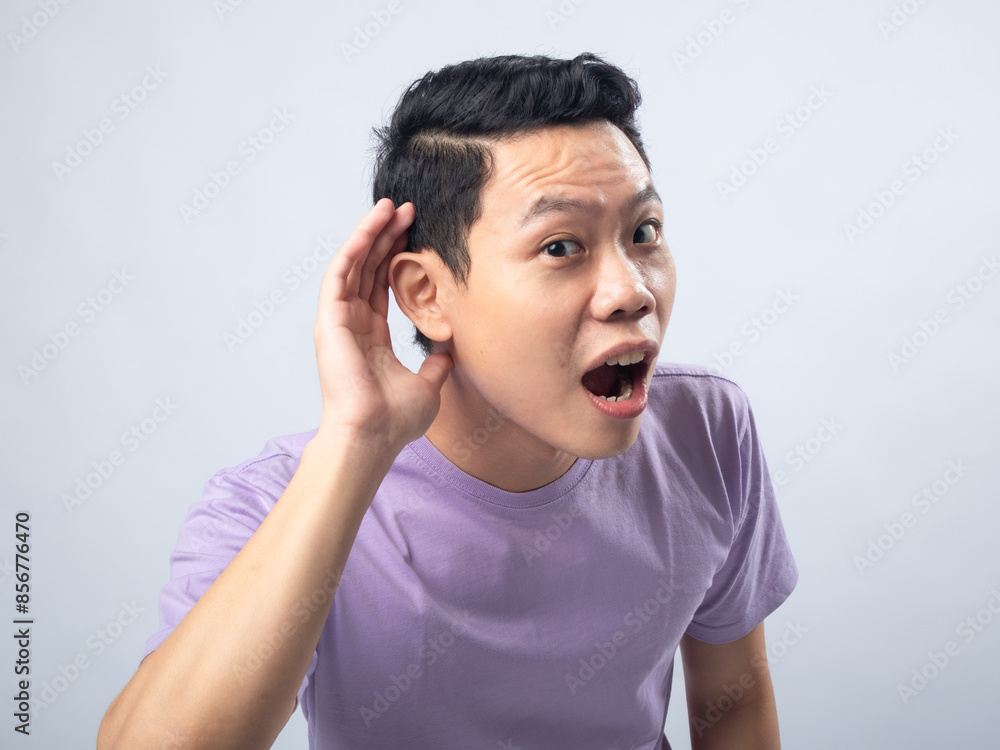 Young Asian man in a lavender t-shirt cupping his ear and looking surprised as if trying to hear something better. Studio shot on a plain background, highlighting his curious expression Young Asian man in a lavender t-shirt cupping his ear and looking surprised as if trying to hear something better. Studio shot on a plain background, highlighting his curious expression