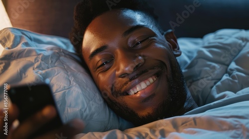 a young smiling african, american man lying in bed, looking at his cell phone
