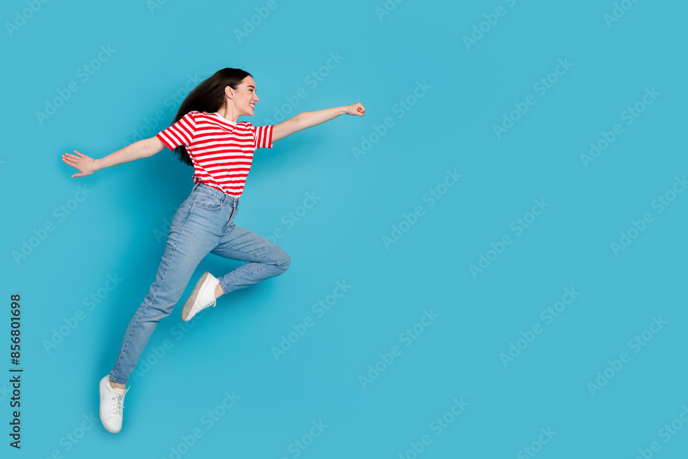 Fototapeta premium Full length photo of adorable lovely girl wear striped t-shirt jumping high rising fist empty space isolated blue color background
