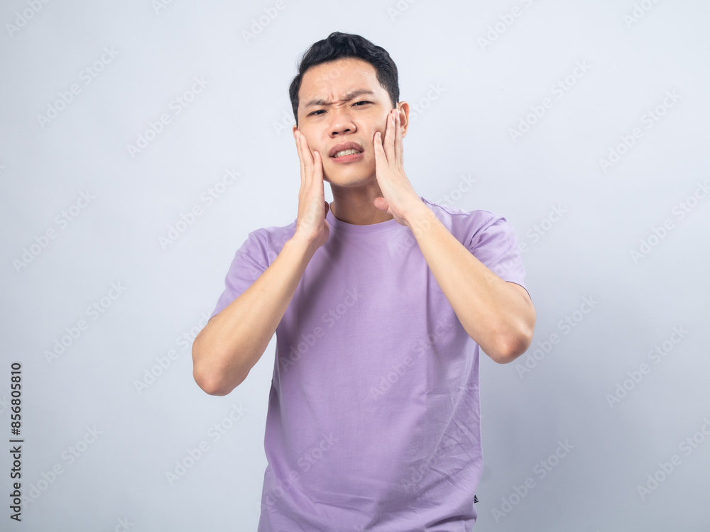 Young Asian man in a lavender t-shirt grimacing in pain while holding his cheek, indicating a toothache. Studio shot on a plain background, highlighting his discomfort and casual fashion. Young Asian man in a lavender t-shirt grimacing in pain while holding his cheek, indicating a toothache. Studio shot on a plain background, highlighting his discomfort and casual fashion.