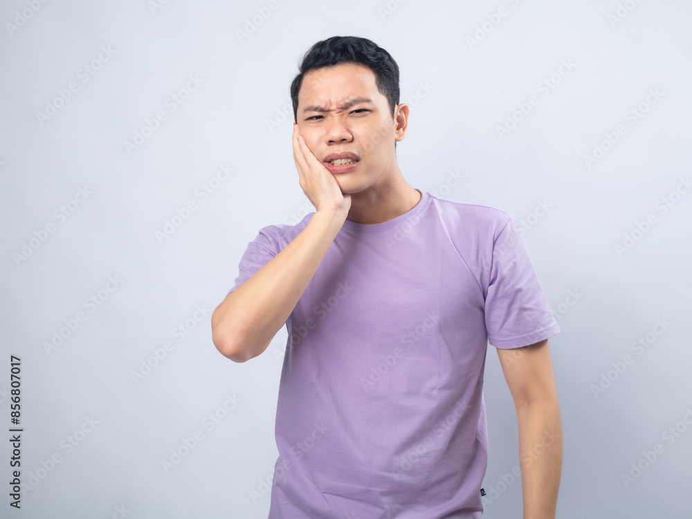 Young Asian man in a lavender t-shirt grimacing in pain while holding his cheek, indicating a toothache. Studio shot on a plain background, highlighting his discomfort and casual fashion. Young Asian man in a lavender t-shirt grimacing in pain while holding his cheek, indicating a toothache. Studio shot on a plain background, highlighting his discomfort and casual fashion.