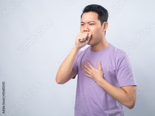 Young Asian man in lavender t-shirt coughing into his fist while holding his chest, indicating discomfort or illness. Studio shot on plain background capturing his casual outfit