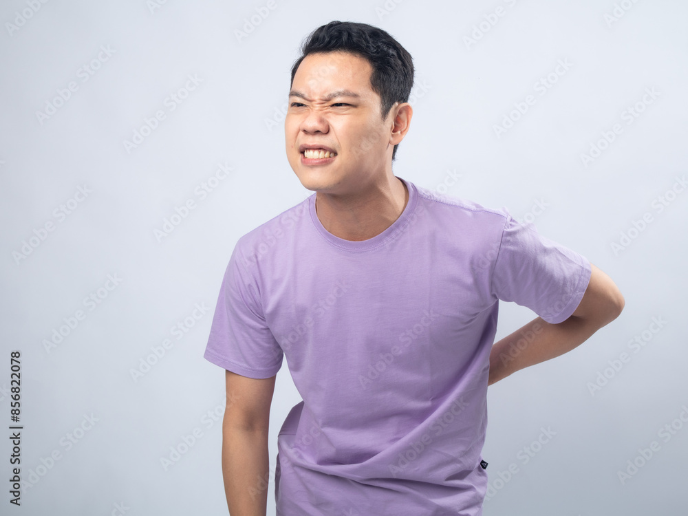 Young Asian man in a lavender t-shirt grimacing in pain and holding his lower back. Studio shot on a plain background, highlighting his discomfort and casual fashion. Young Asian man in a lavender t-shirt grimacing in pain and holding his lower back. Studio shot on a plain background, highlighting his discomfort and casual fashion.