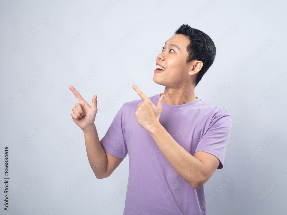 Young Asian man in a lavender t-shirt pointing with both hands and looking excitedly in the direction he's pointing. Studio shot on a plain background Young Asian man in a lavender t-shirt pointing with both hands and looking excitedly in the direction he's pointing. Studio shot on a plain background