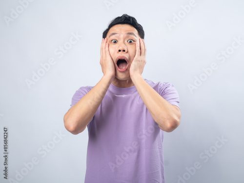 Young Asian man in a lavender t-shirt holding his face in shock with wide eyes and mouth open. Studio shot on a plain background, emphasizing his surprised expression and casual fashion.