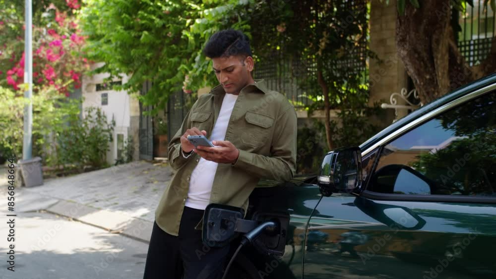 Medium long shot of busy Indian man with smartphone leaning on electric car charging with cable outdoors Medium long shot of busy Indian man with smartphone leaning on electric car charging with cable outdoors