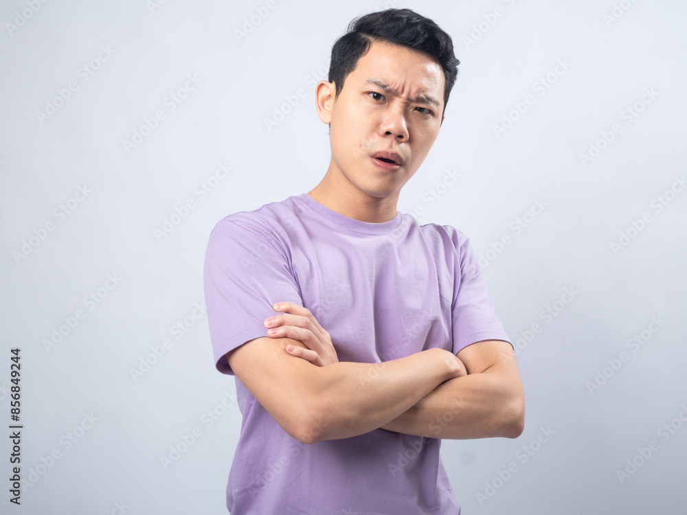 Obraz premium Young Asian man in a lavender t-shirt standing with arms crossed, looking serious and thoughtful. Studio shot on a plain background emphasizing his focused expression and casual attire