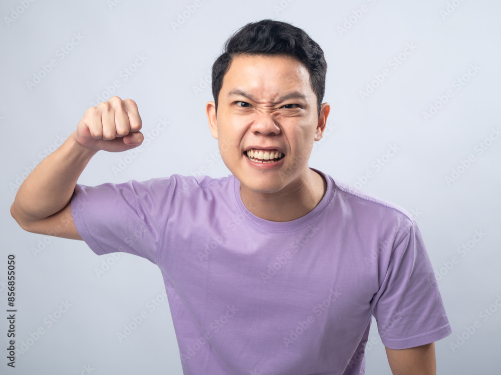 Young Asian man in a lavender t-shirt with an angry expression, clenching his fist and showing aggression. Studio shot on a plain background, highlighting his intense emotion and casual fashion. Young Asian man in a lavender t-shirt with an angry expression, clenching his fist and showing aggression. Studio shot on a plain background, highlighting his intense emotion and casual fashion.