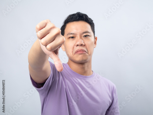 Young Asian man in a lavender t-shirt showing a disapproving expression and giving a thumbs-down gesture. Studio shot on a plain background, highlighting his displeased expression photo