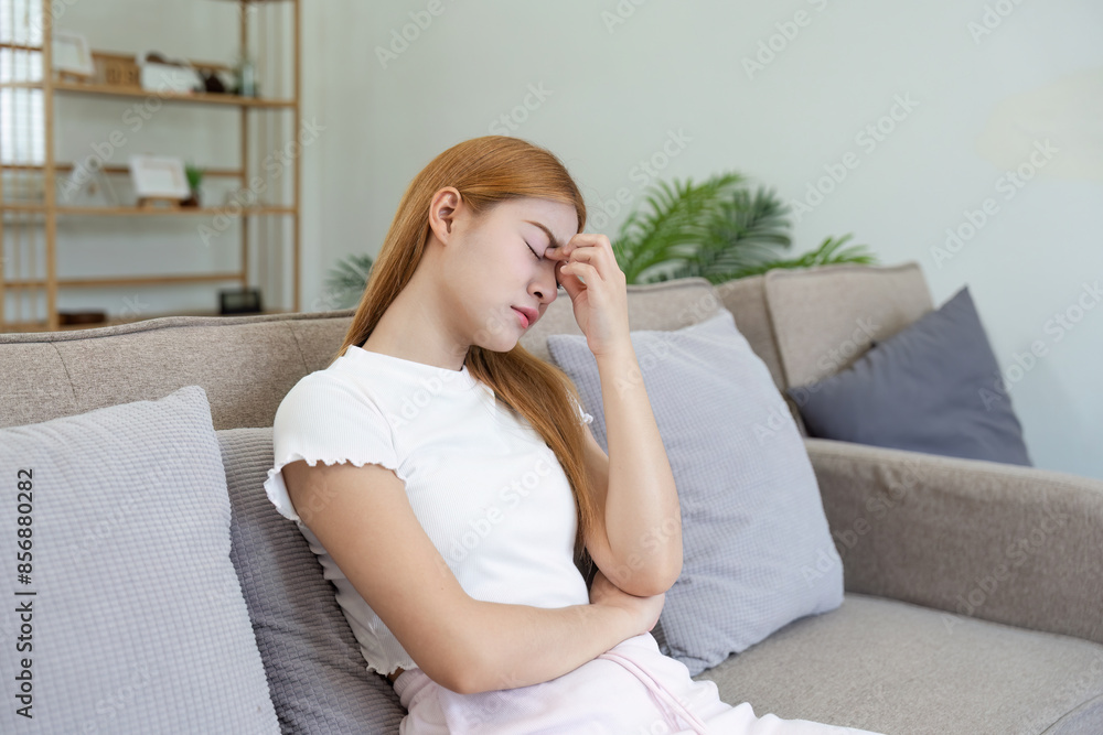 Young Woman Sitting on Sofa Holding Her Head in Pain Due to a Headache in a Modern Living Room Setting Young Woman Sitting on Sofa Holding Her Head in Pain Due to a Headache in a Modern Living Room Setting