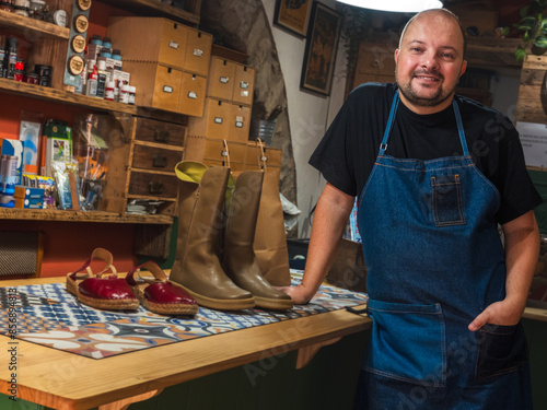 Φωτογραφία Shoemaker poses in front of the camera next to the counter of his store with different repaired shoes