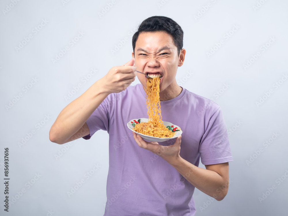 Young Asian man in a lavender t-shirt enthusiastically eating instant noodles from a bowl with a fork. Studio shot on a plain background, highlighting his joyful expression and casual fashion Young Asian man in a lavender t-shirt enthusiastically eating instant noodles from a bowl with a fork. Studio shot on a plain background, highlighting his joyful expression and casual fashion