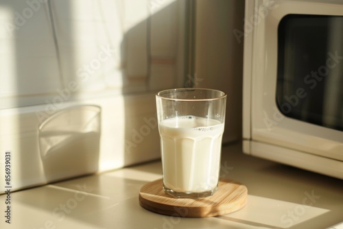 A glass of milk is placed on a wooden coaster, with a microwave behind and a light white beige wall in front.