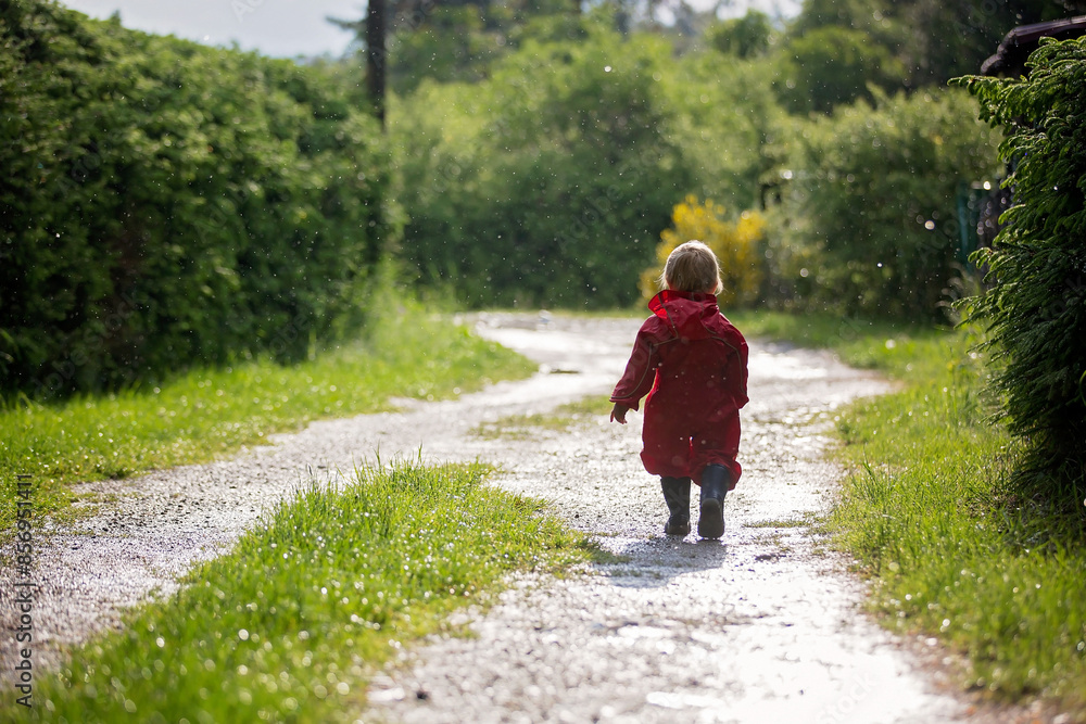 Sweet little toddler boy, blond child in red raincoat and blue boots, playing in the rain in muddy puddles, jumping and laughing Sweet little toddler boy, blond child in red raincoat and blue boots, playing in the rain in muddy puddles, jumping and laughing