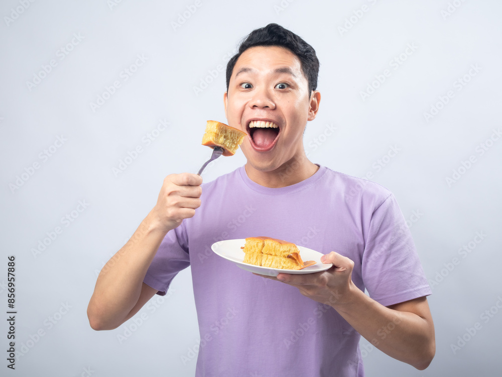 Young Asian man in a lavender t-shirt excitedly holding a plate with a slice of cake on a fork. Studio shot on a plain background, highlighting his joyful expression and casual fashion.