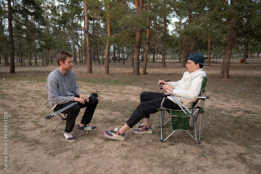 Two men sit on folding chairs in a forest clearing during a sunny afternoon. They are enjoying a picnic in the shade of the trees, with a peaceful and serene atmosphere Two men sit on folding chairs in a forest clearing during a sunny afternoon. They are enjoying a picnic in the shade of the trees, with a peaceful and serene atmosphere