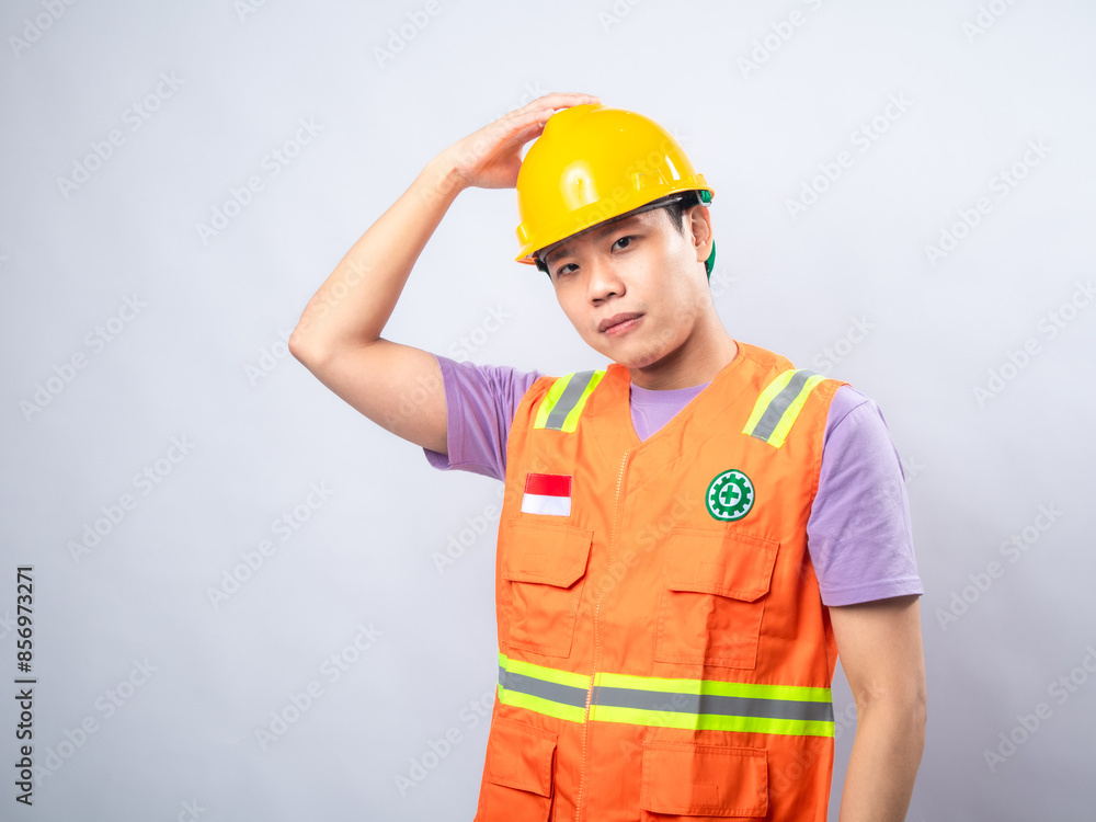 An Asian male construction worker, wearing an orange safety vest and a yellow hard hat, touches his head with a thoughtful expression. The plain white background highlights the worker An Asian male construction worker, wearing an orange safety vest and a yellow hard hat, touches his head with a thoughtful expression. The plain white background highlights the worker