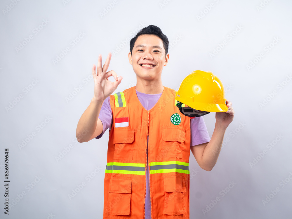 An Asian male construction worker, wearing an orange safety vest and holding a yellow hard hat, smiles and gives an An Asian male construction worker, wearing an orange safety vest and holding a yellow hard hat, smiles and gives an