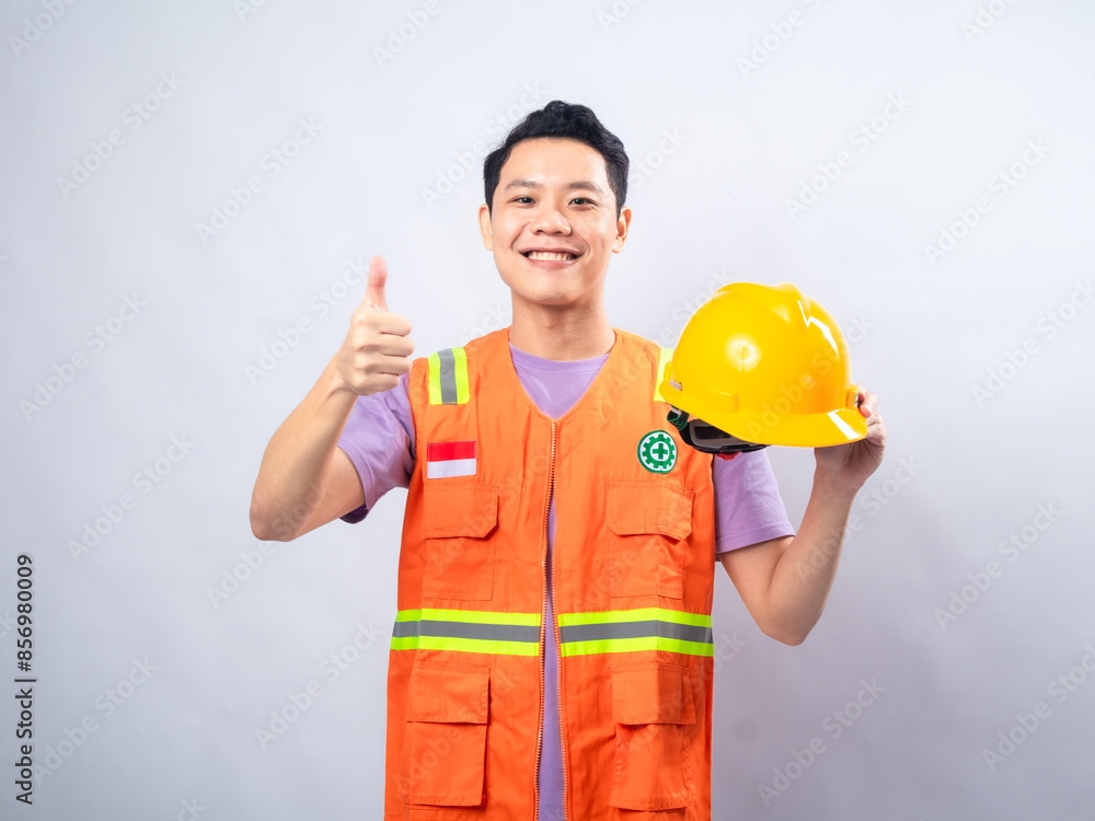 An Asian male construction worker, wearing an orange safety vest and holding a yellow hard hat, smiles and gives an An Asian male construction worker, wearing an orange safety vest and holding a yellow hard hat, smiles and gives an