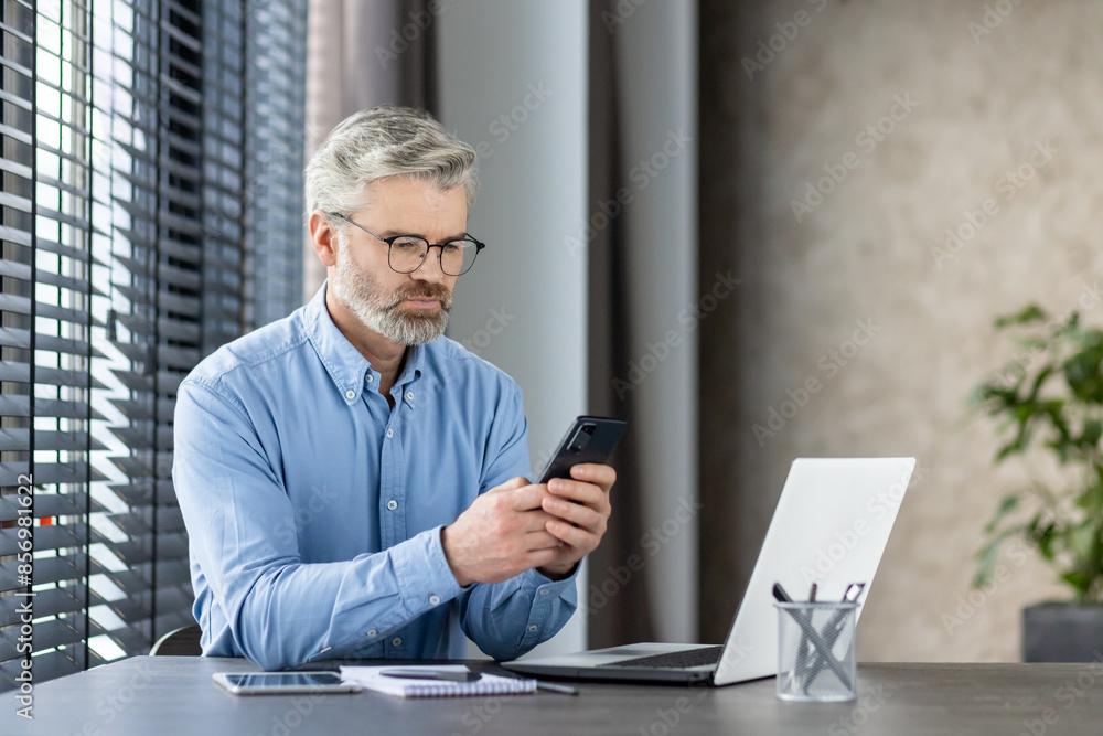 Mature businessman sitting at a desk, attentively using a mobile phone and working on a laptop in a modern office environment. Mature businessman sitting at a desk, attentively using a mobile phone and working on a laptop in a modern office environment.