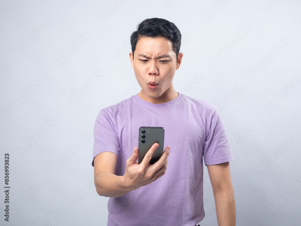 Young Asian man in a lavender t-shirt looking surprised and excited while waving at his smartphone. Studio shot on a plain background, highlighting his enthusiastic expression and casual fashion. Young Asian man in a lavender t-shirt looking surprised and excited while waving at his smartphone. Studio shot on a plain background, highlighting his enthusiastic expression and casual fashion.