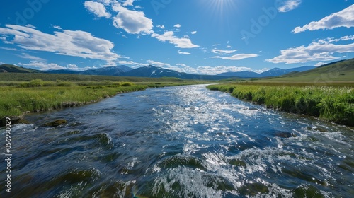 Mountain river flowing under a clear blue sky, surrounded by green meadows and distant mountains, creating a serene and picturesque landscape.	