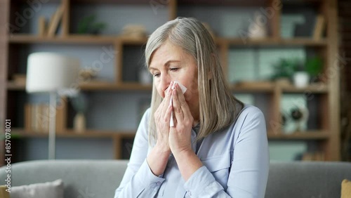 Senior gray haired female with an allergy sneezes sitting on sofa in living room at home. Sick allergic elderly retired woman wipes her nose with a handkerchief. She has a cold, virus or flu. Close up photo